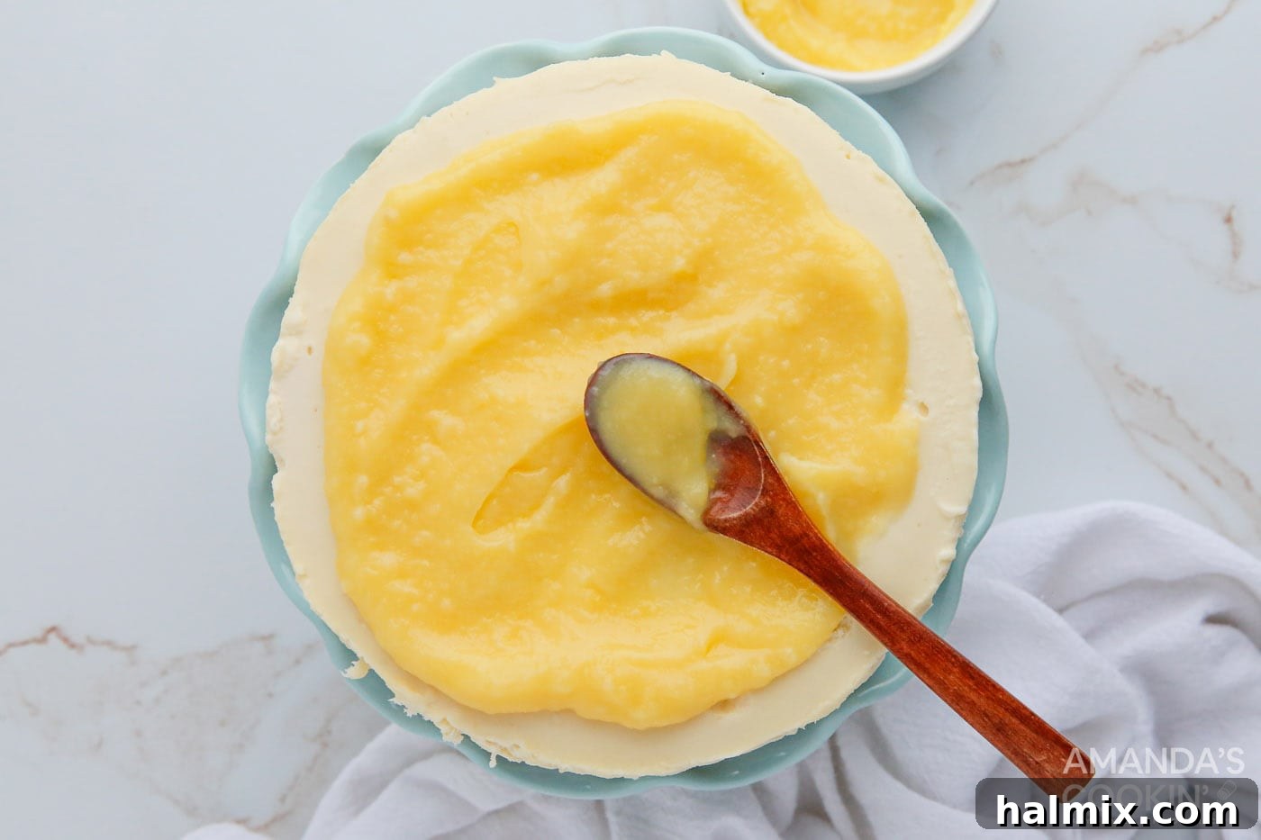 A spoon delicately spreads a layer of smooth, yellow lemon curd over the top of the chilled cheesecake, preparing it for serving.