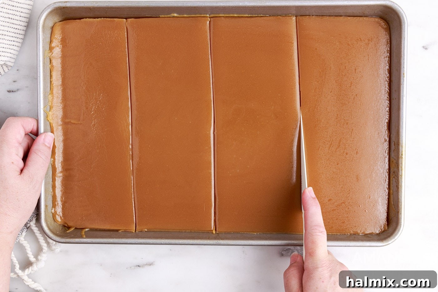 A butter knife slicing through a sheet of set microwave caramels on a parchment-lined pan.