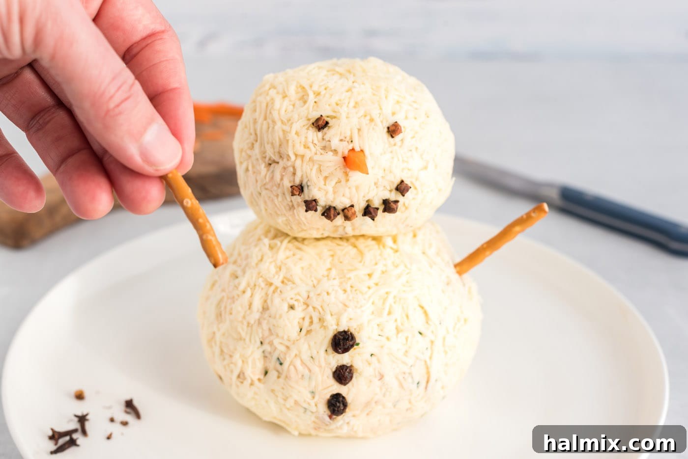 Festive Snowman Cheeseball 12 hand adding a pretzel stick to snowman cheeseball