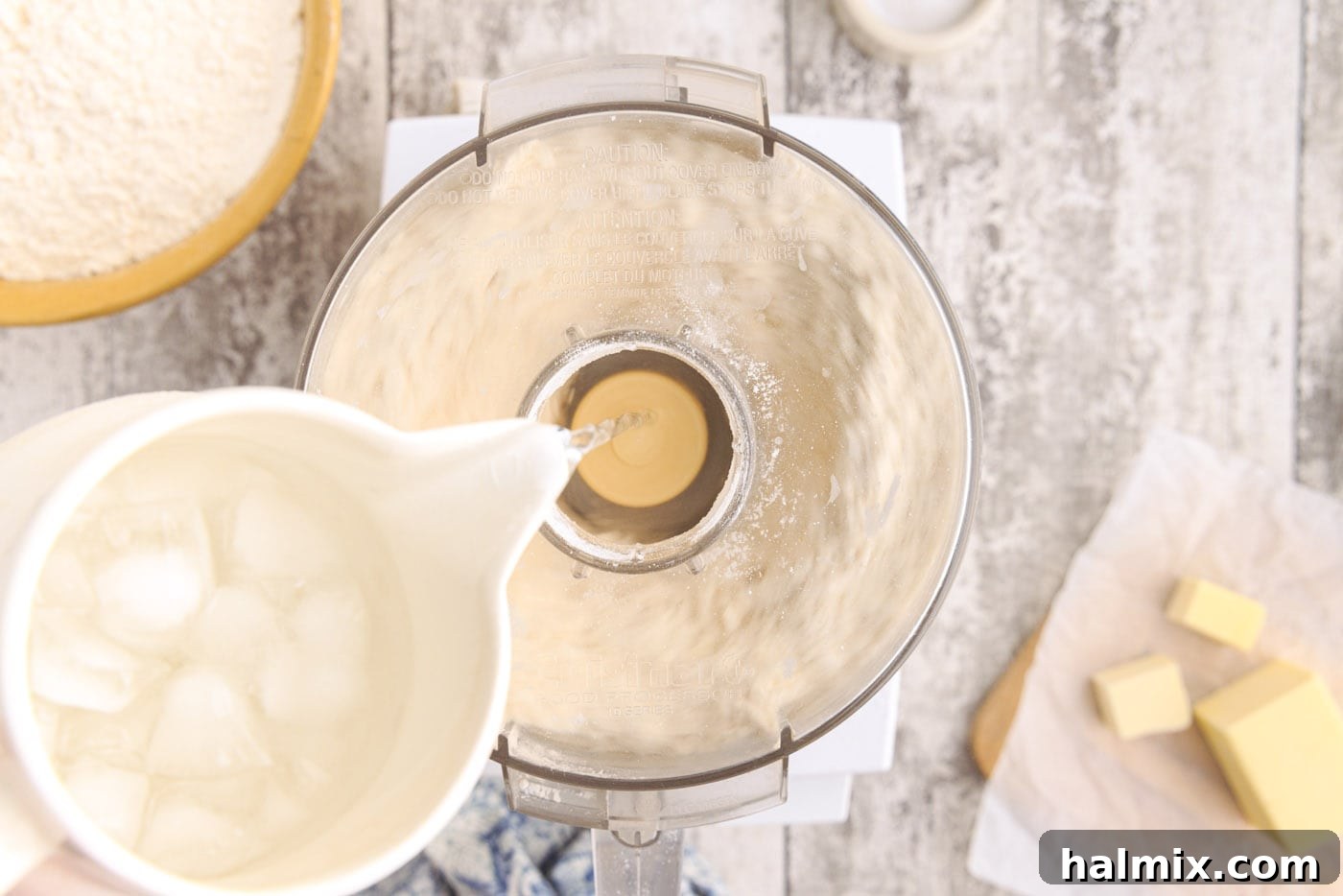 Ice water being drizzled into a food processor with butter and flour mixture