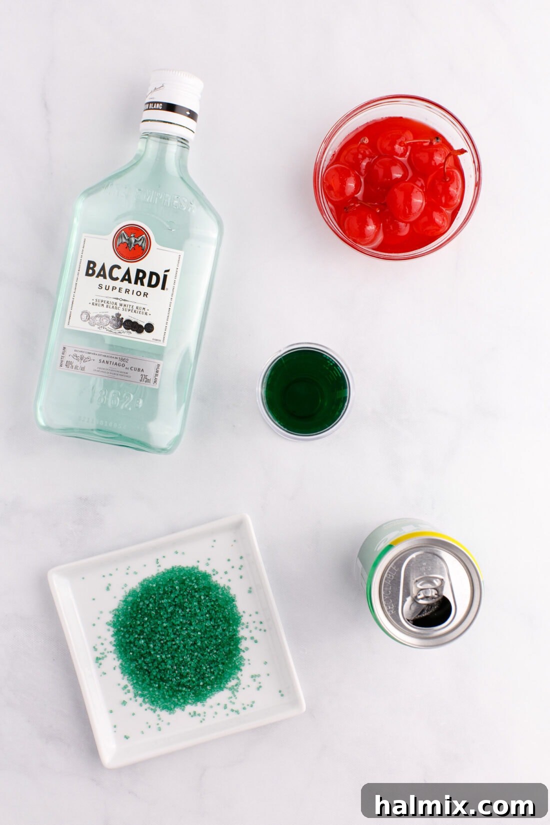 A flat lay showing all ingredients for the Grinch Cocktail: Midori, white rum, lemon-lime soda, maraschino cherries, lime, and green sugar.