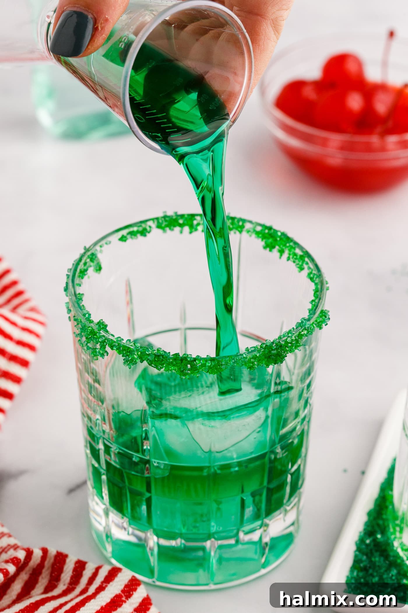 Close-up image of a hand pouring Midori into a rock glass filled with ice, showing the start of the Grinch Cocktail preparation.