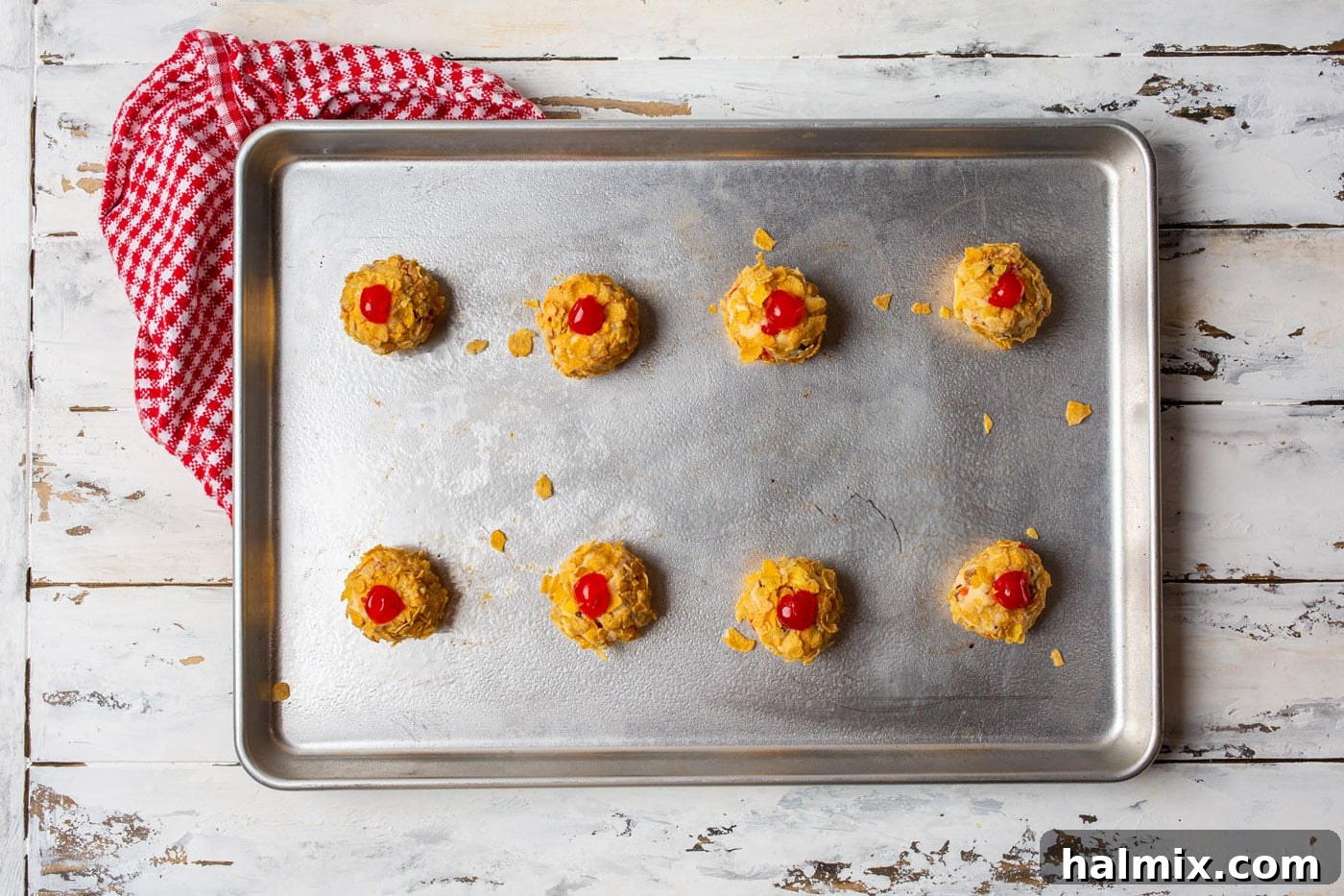 Cherry wink cookie dough balls coated in cornflakes and topped with extra cherry halves, neatly arranged on a baking sheet before baking.