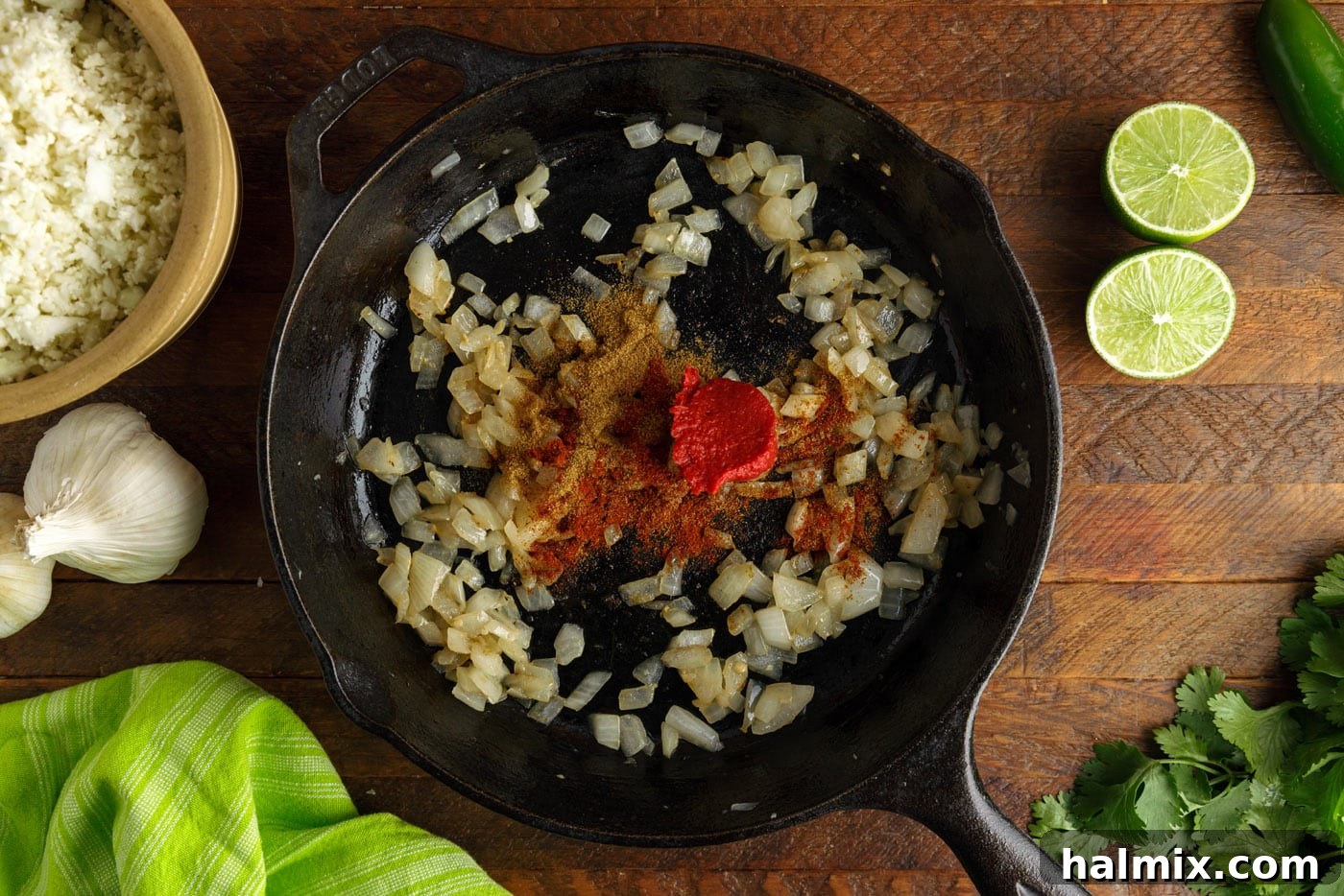 Tomato paste and spices (cumin, chili powder) being mixed into sautéed onion and garlic in a skillet.