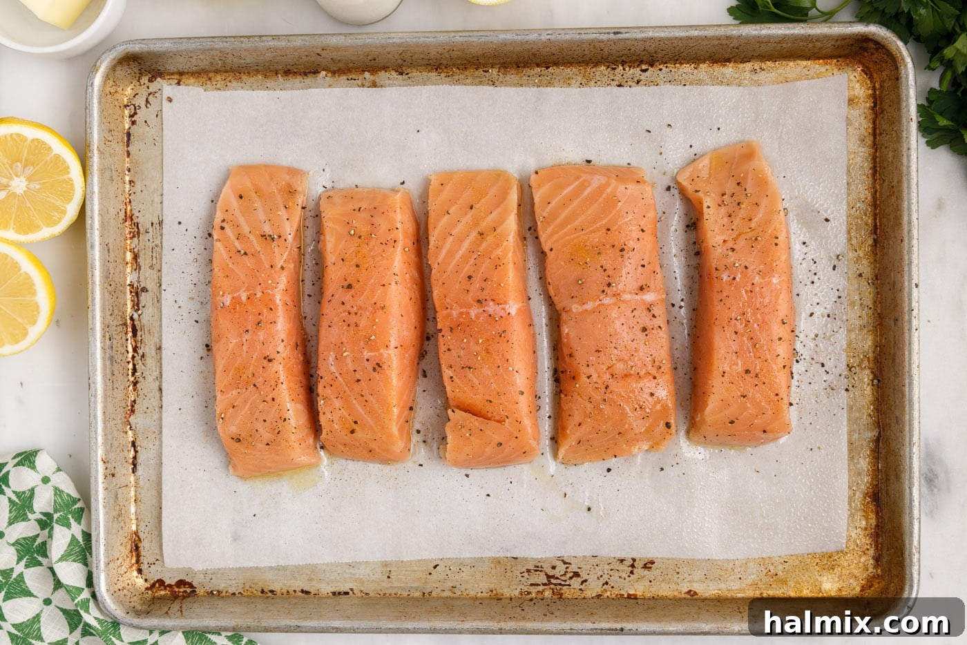Seasoned salmon fillets, dusted with black pepper and seasoned salt, on a baking sheet.