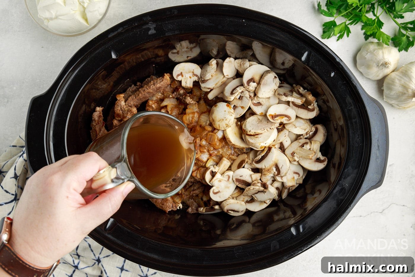 pouring beef stock into crockpot with mushrooms,, onions, and steak