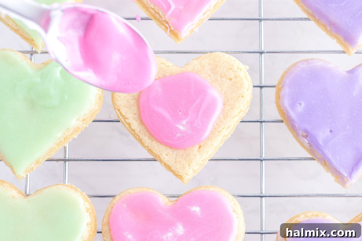 A spoon dolloping pink frosting onto a heart-shaped sugar cookie