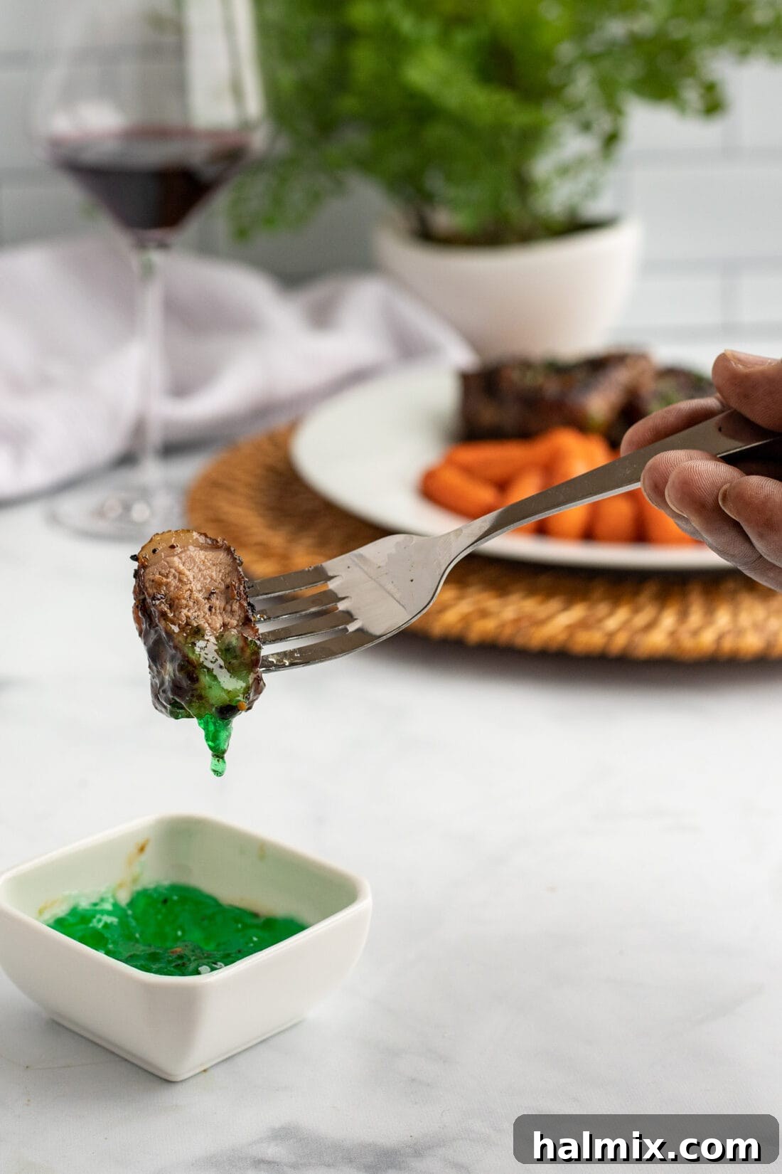 A fork holding a piece of lamb chop, dipping it into a small dish of mint jelly.