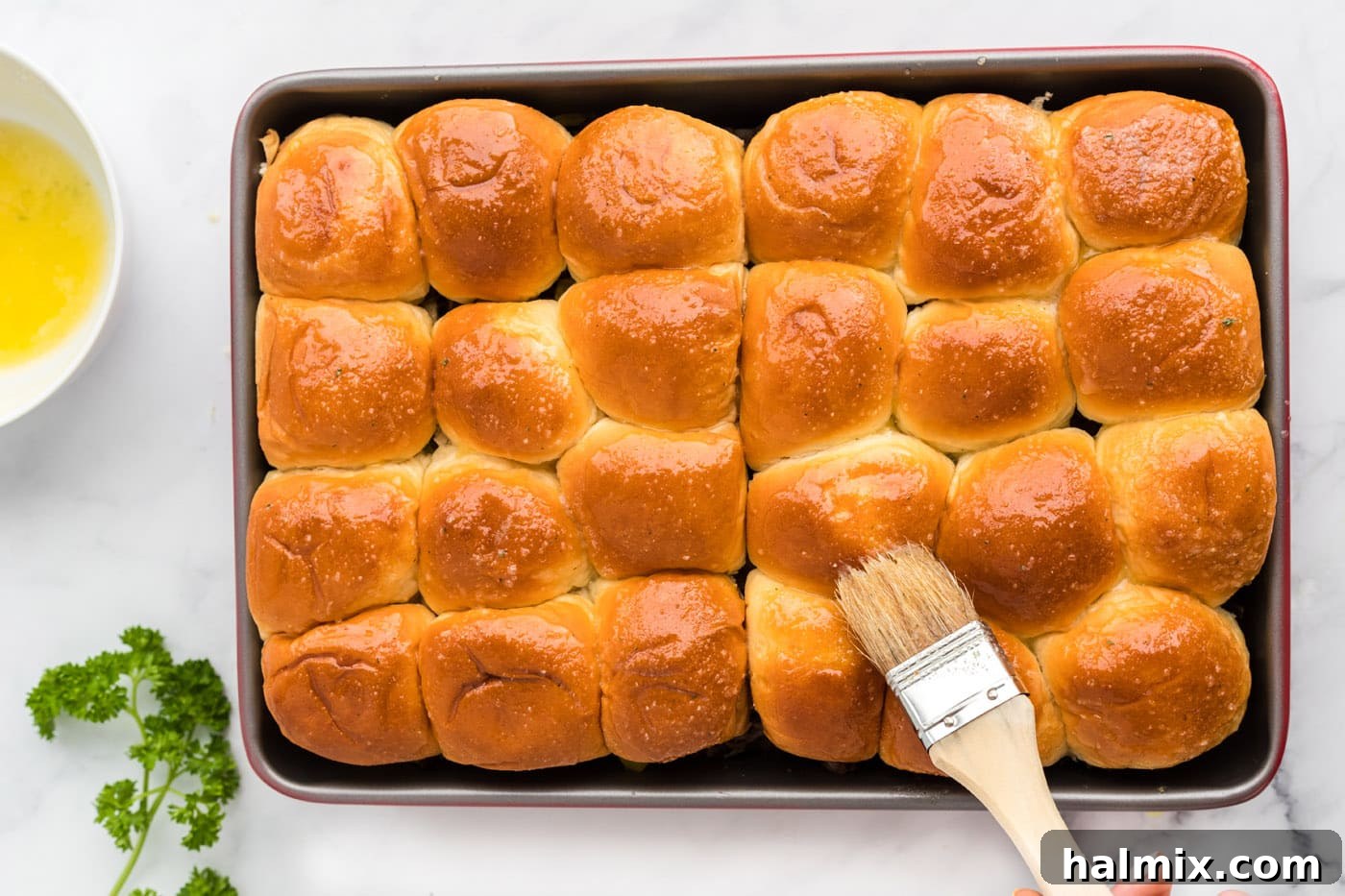 A pastry brush applying melted garlic butter onto the tops of the Mississippi Pot Roast Sliders in the pan.