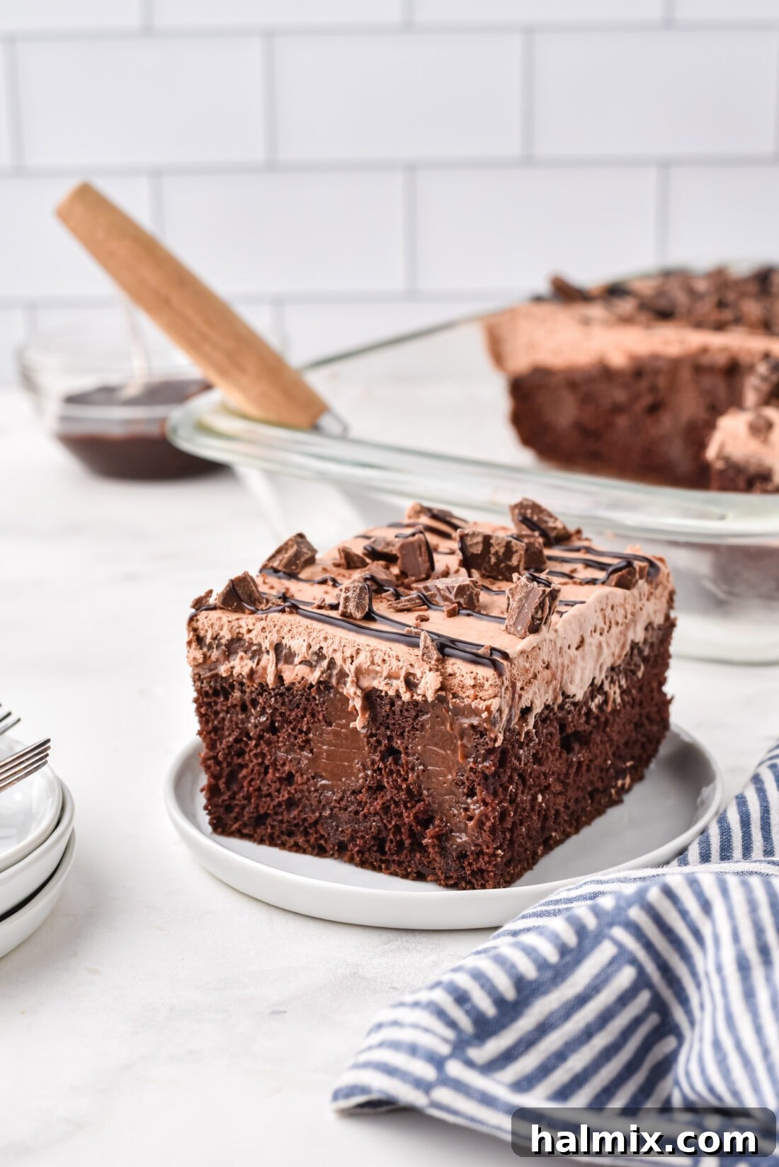 A perfectly sliced piece of moist Chocolate Poke Cake on a white plate, showcasing its layers of rich chocolate cake, pudding, and whipped topping, garnished with chocolate syrup and candy bits.