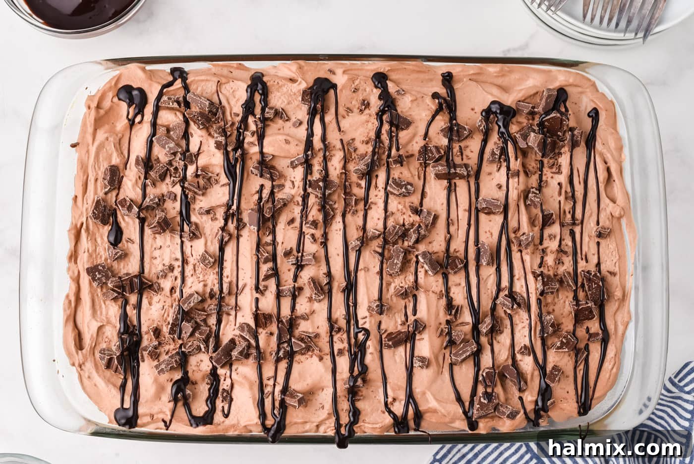 A fully assembled Triple Chocolate Poke Cake garnished with chopped chocolate candy and a rich chocolate syrup drizzle, ready to be chilled.