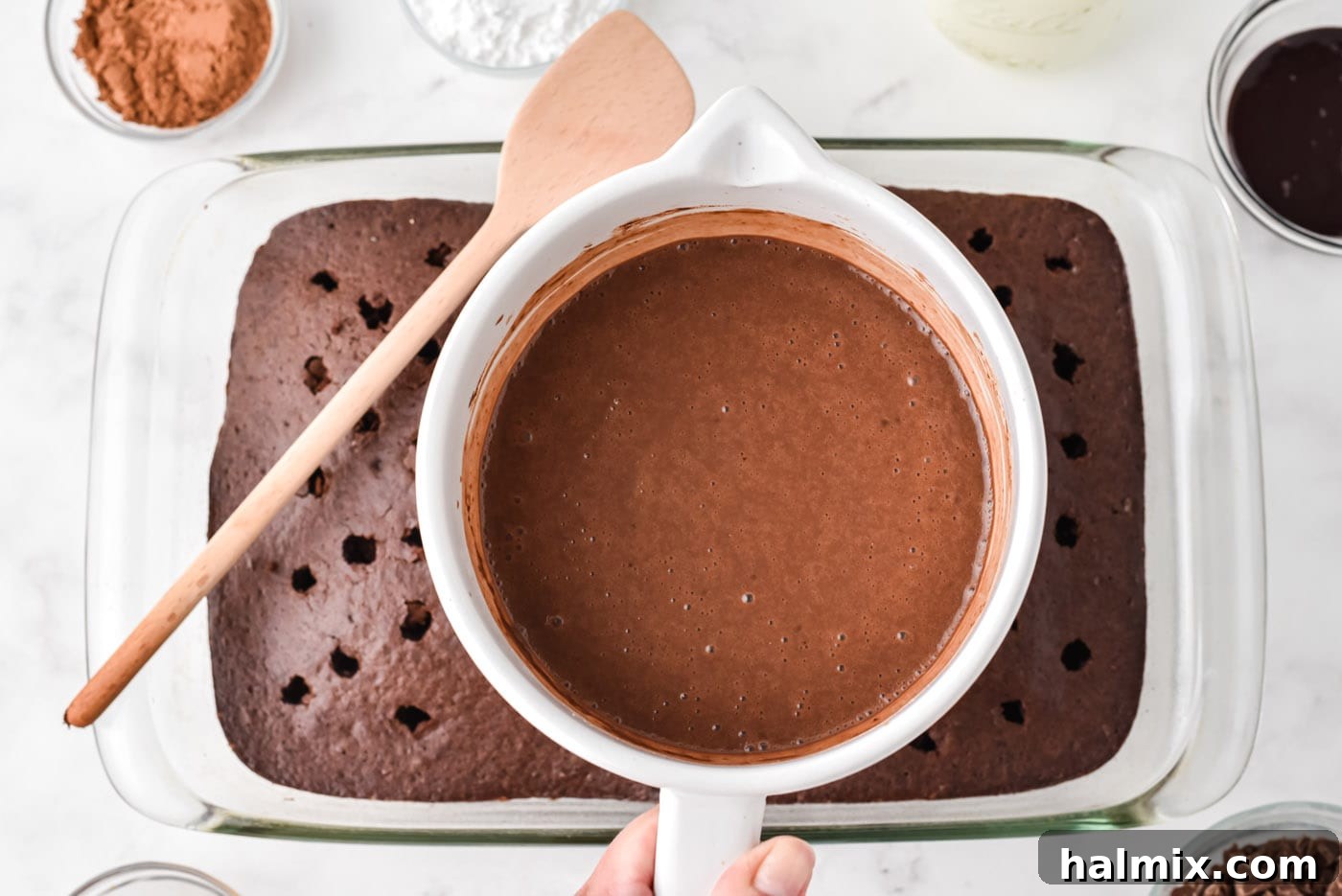 Thick chocolate pudding being poured generously over the surface of the warm, poked chocolate cake in a baking dish.