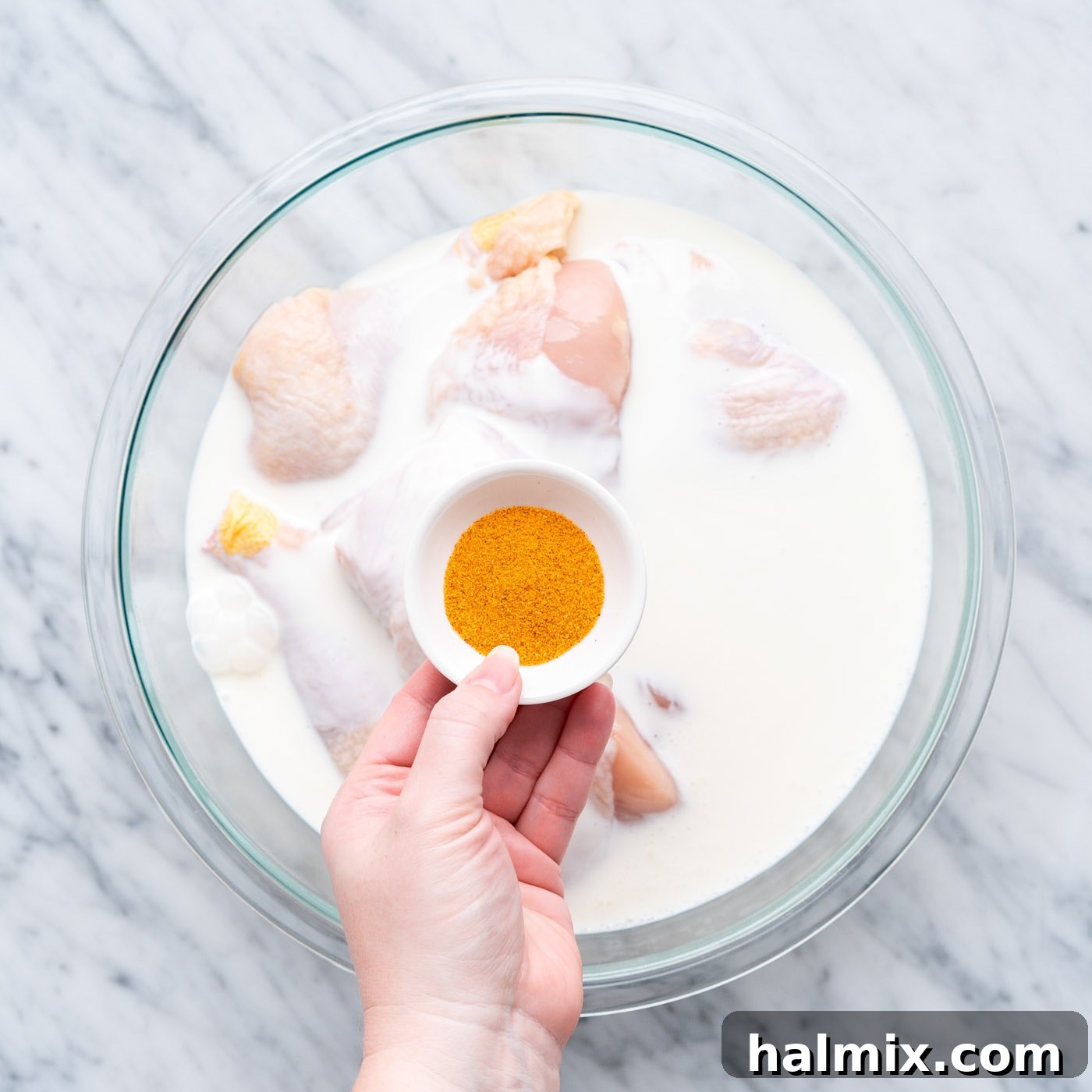 Buttermilk and seasoned salt being poured over chicken pieces in a large bowl, demonstrating the start of the marinating process.