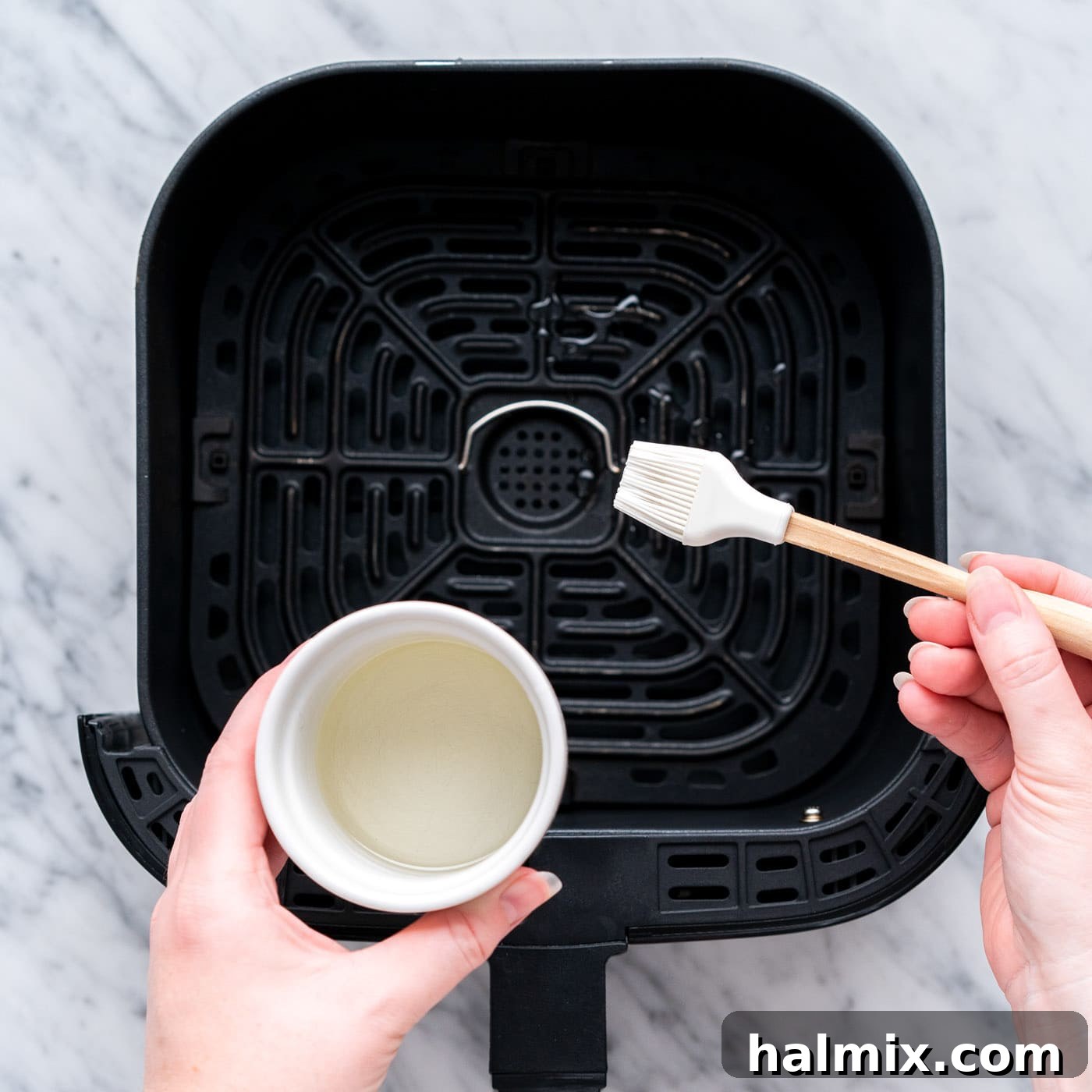 A hand brushing a small amount of vegetable oil onto the bottom of an air fryer basket, preparing it for cooking and preventing chicken from sticking.