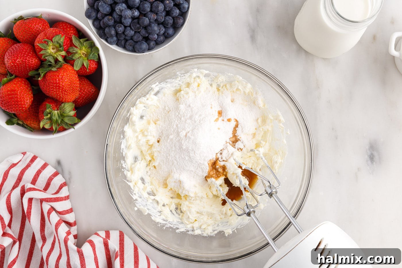 Cream cheese, pudding mix, milk, and vanilla extract being combined in a bowl with a mixer.