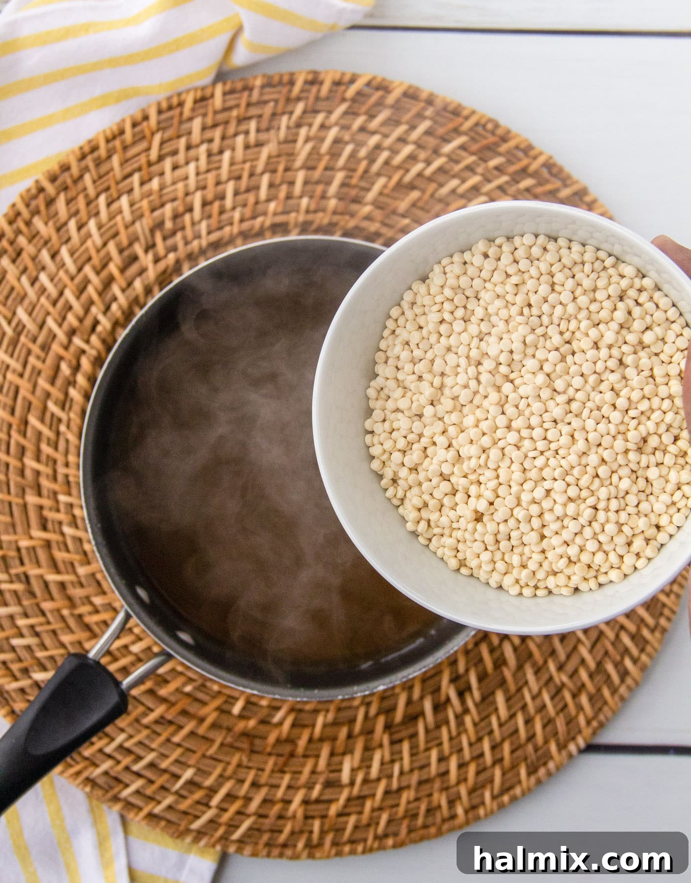 Pearl couscous being poured into a saucepan of boiling broth on a stovetop.