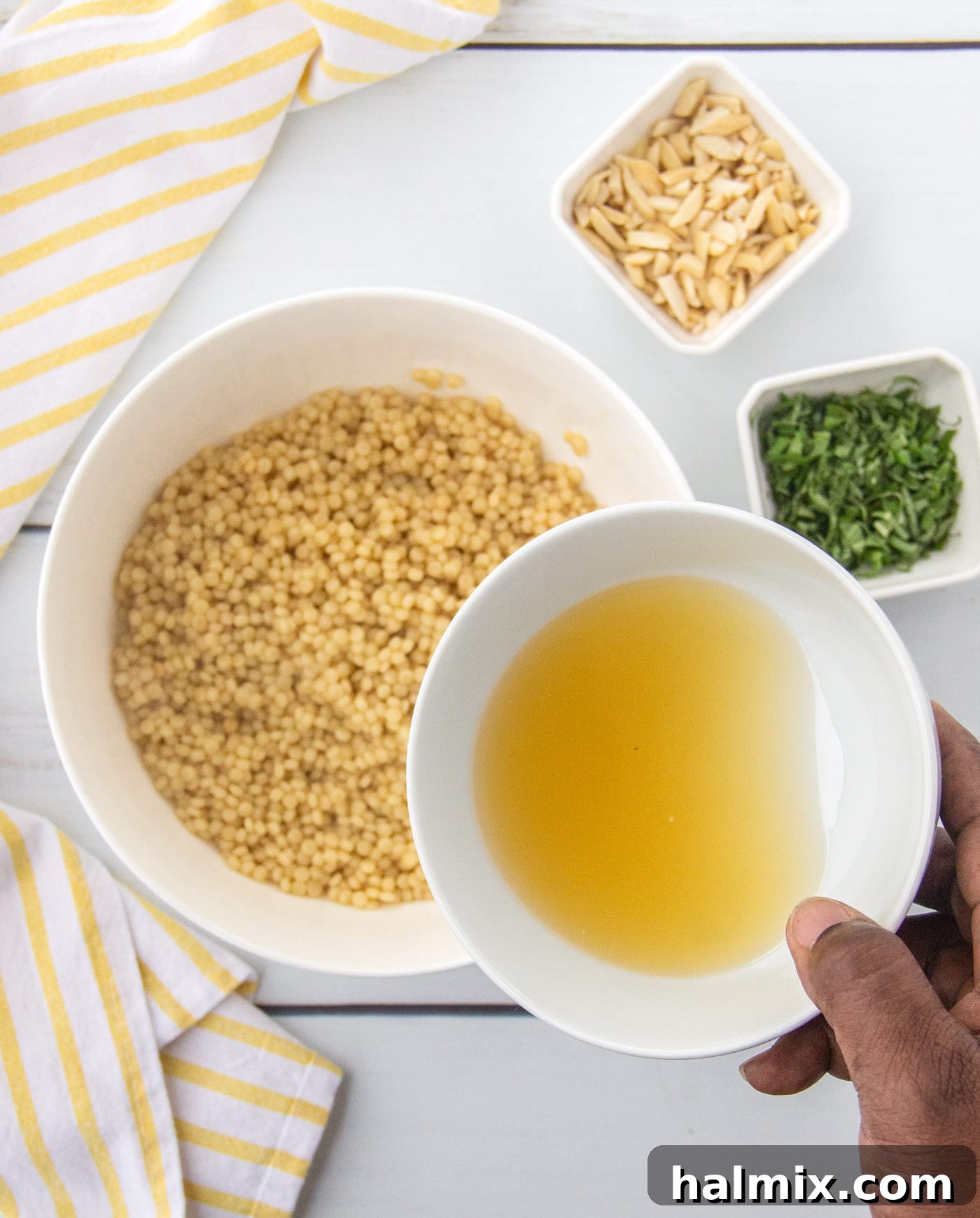 Lemon juice mixture, sliced basil, and slivered almonds being added to cooked couscous in a large mixing bowl, ready for stirring.