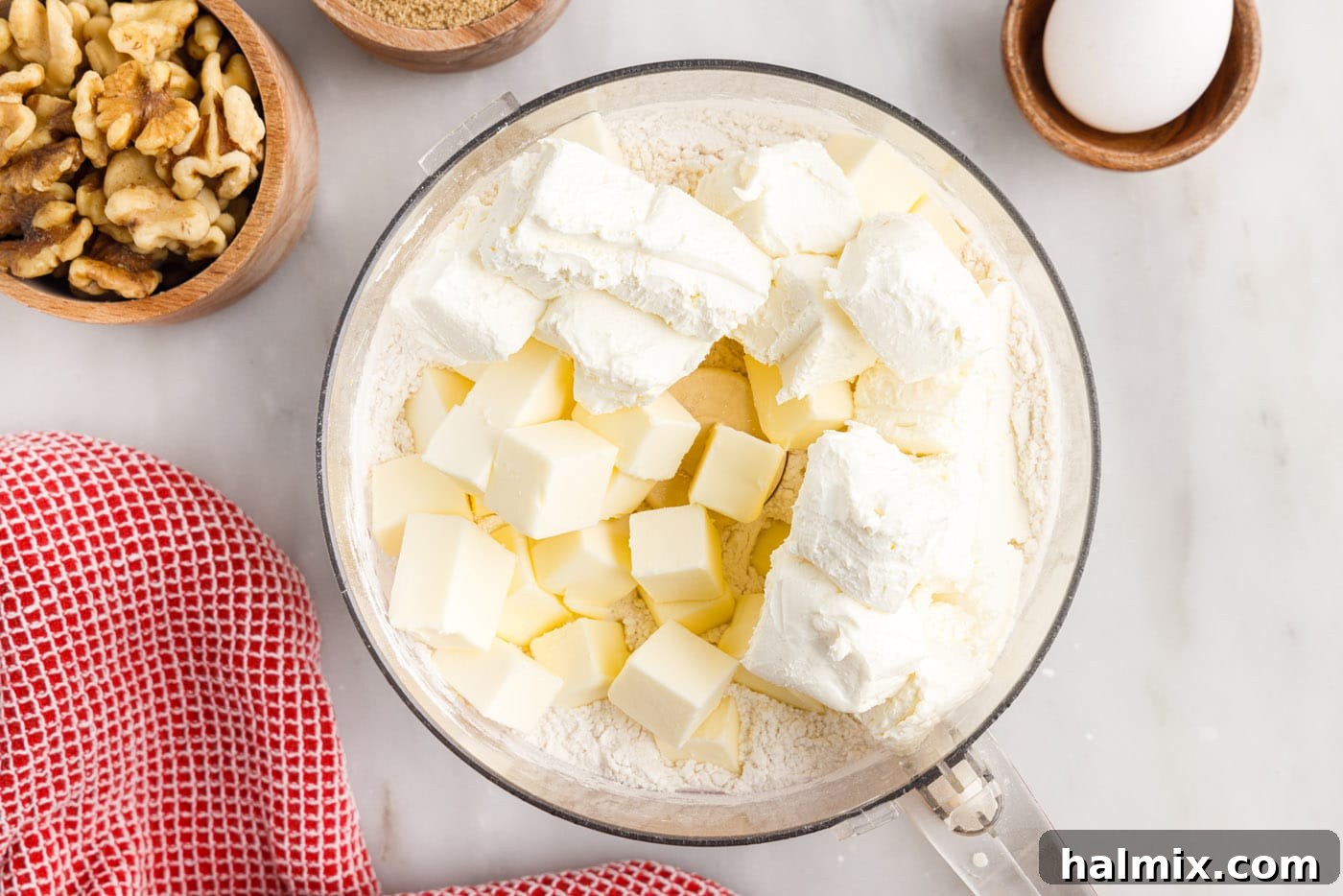 Cold cream cheese and butter cubes are added over flour in a food processor bowl, ready to be pulsed.
