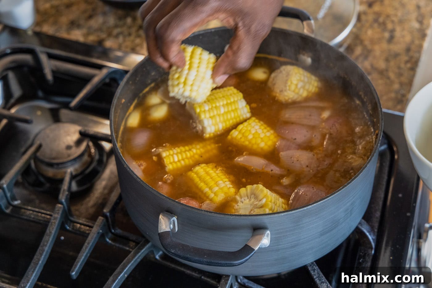 A hand carefully adding pieces of fresh corn on the cob to the simmering shrimp boil pot.
