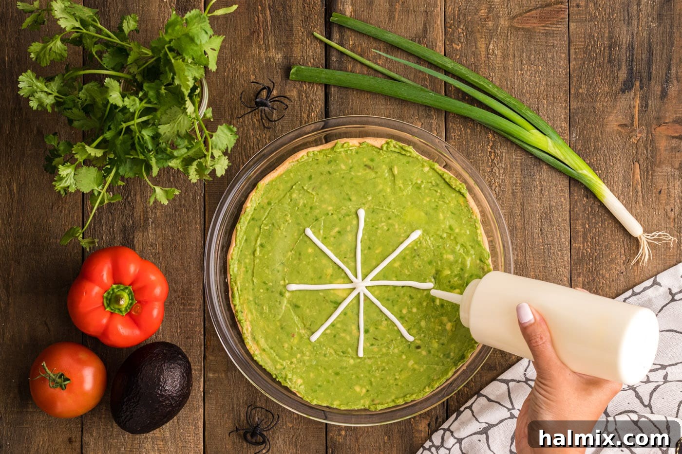 A hand expertly piping sour cream in a concentric circular pattern onto the guacamole layer, forming the base of the spider web.