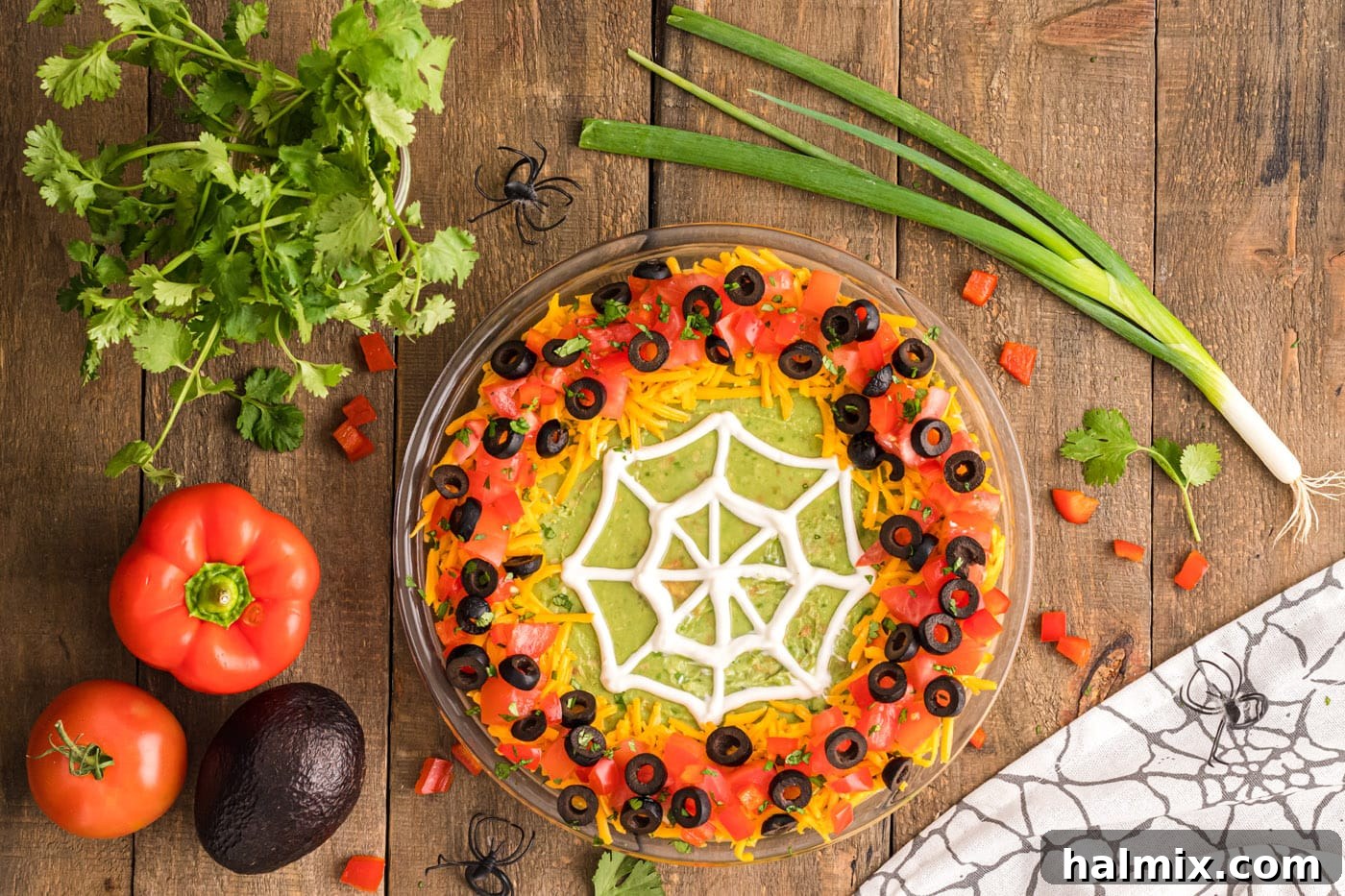An overhead view of the finished Spider Web Taco Dip, garnished with diced tomatoes, sliced black olives, and cilantro around the cheesy border, with the sour cream web in the center.