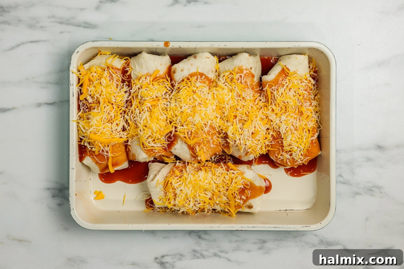 Enchiritos in a baking pan, generously topped with warm enchilada sauce and shredded cheddar cheese, ready for baking.
