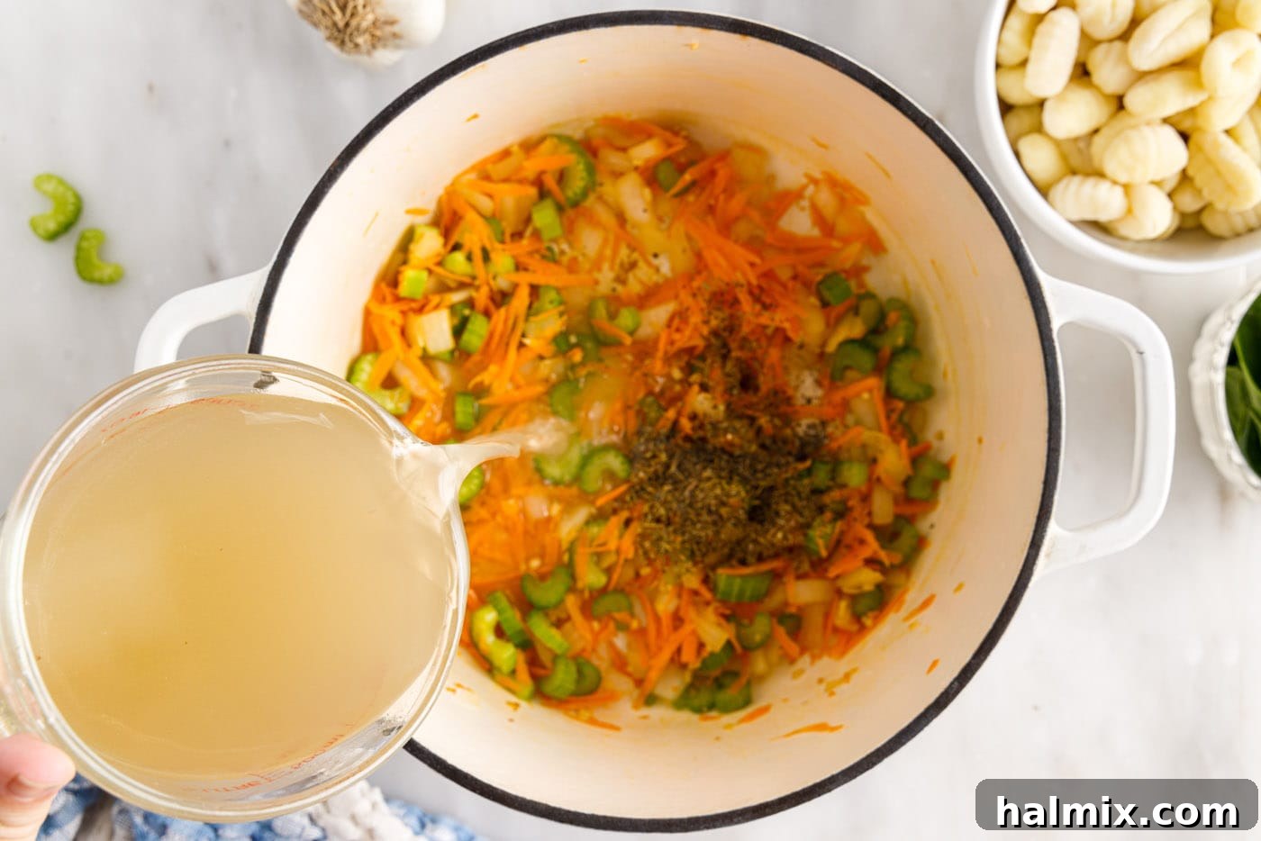 Pouring rich chicken broth over the sautéed vegetables in a large pot with herbs and spices.
