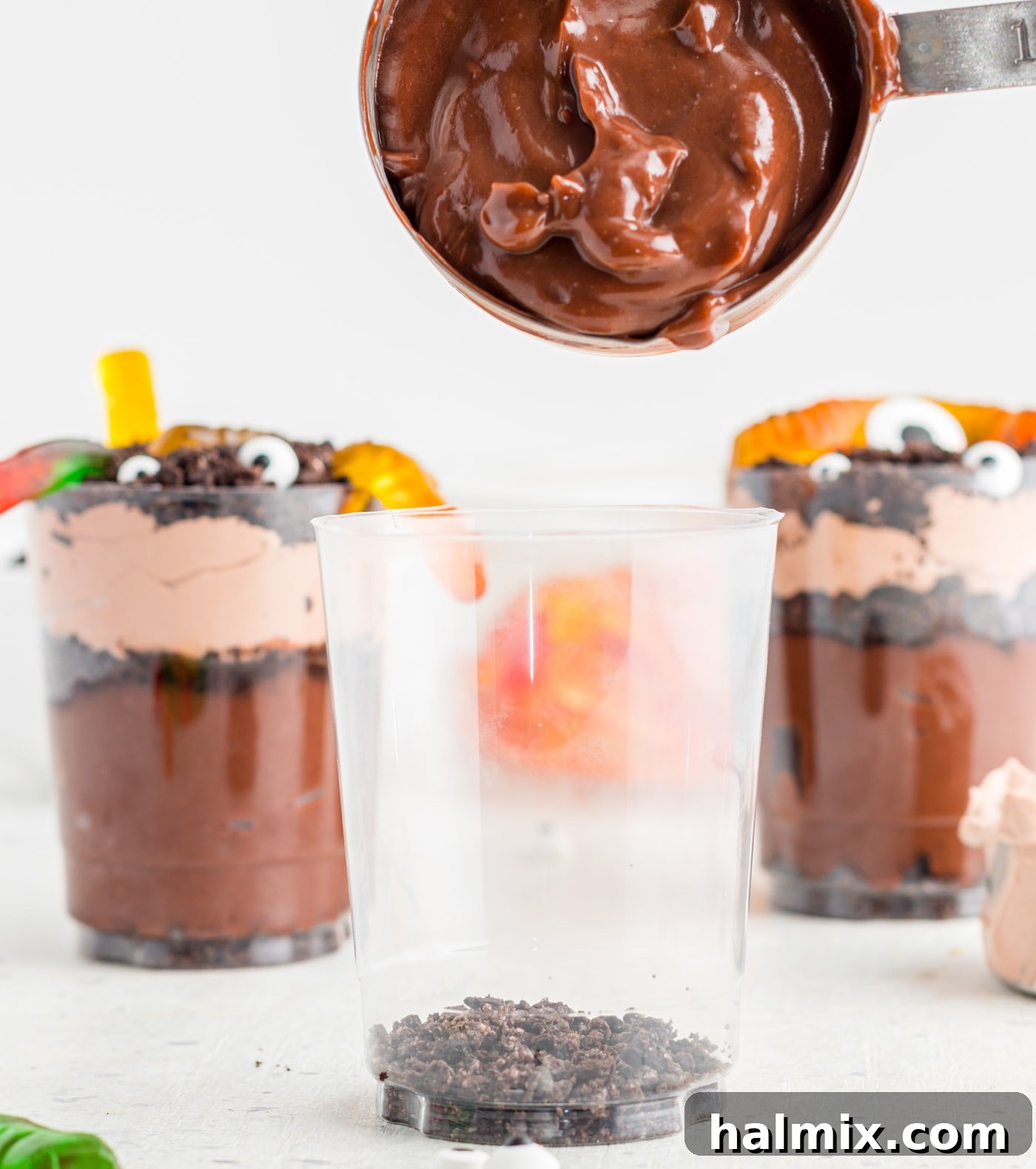 A tablespoon of chocolate pudding being carefully added as a layer over crushed Oreos in a transparent dirt cup.