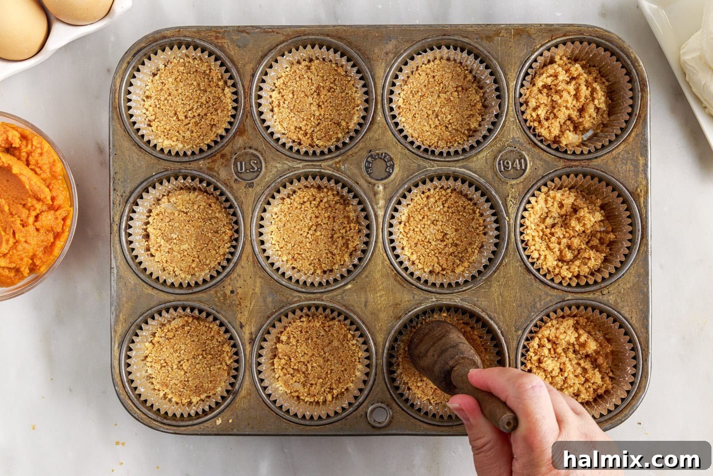 Spiced Pumpkin Cheesecake Minis 7 A hand pressing the graham cracker crust mixture into a muffin tin lined with cupcake wrappers.