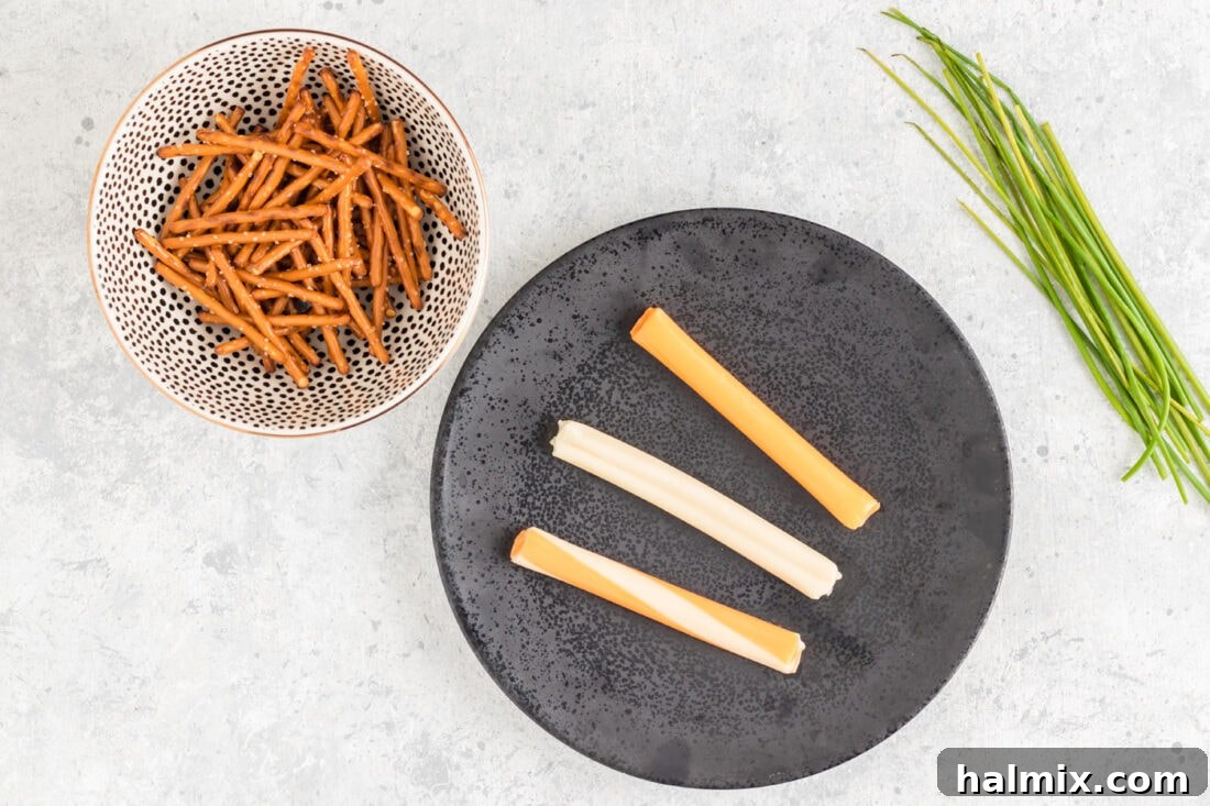 Cheesy Pretzel Broomsticks 4 The three core ingredients for Cheese and Pretzel Broomsticks: string cheese, pretzel sticks, and fresh chives, laid out on a cutting board.