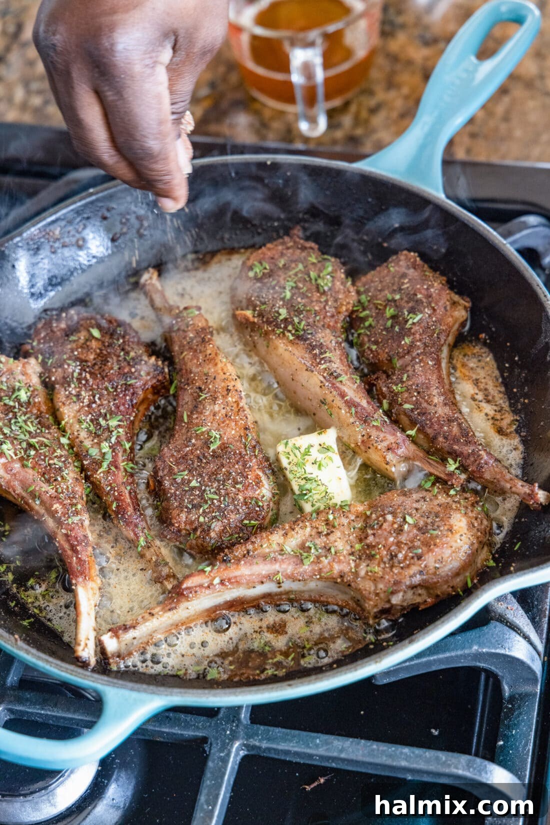 Close-up of a hand sprinkling fresh thyme over sizzling Lollipop Lamb Chops in a skillet, highlighting the golden crust and aromatic herbs.