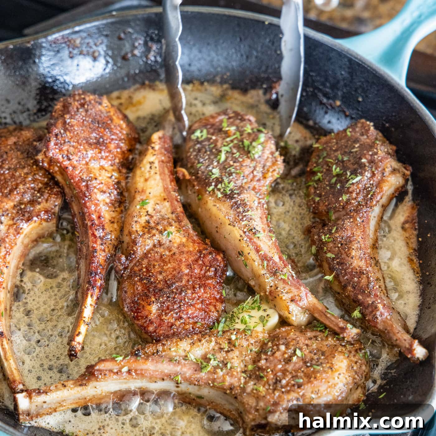 A kitchen tongs turning a lollipop lamb chop in a skillet, ensuring it's fully coated in the glistening butter and herb mixture.