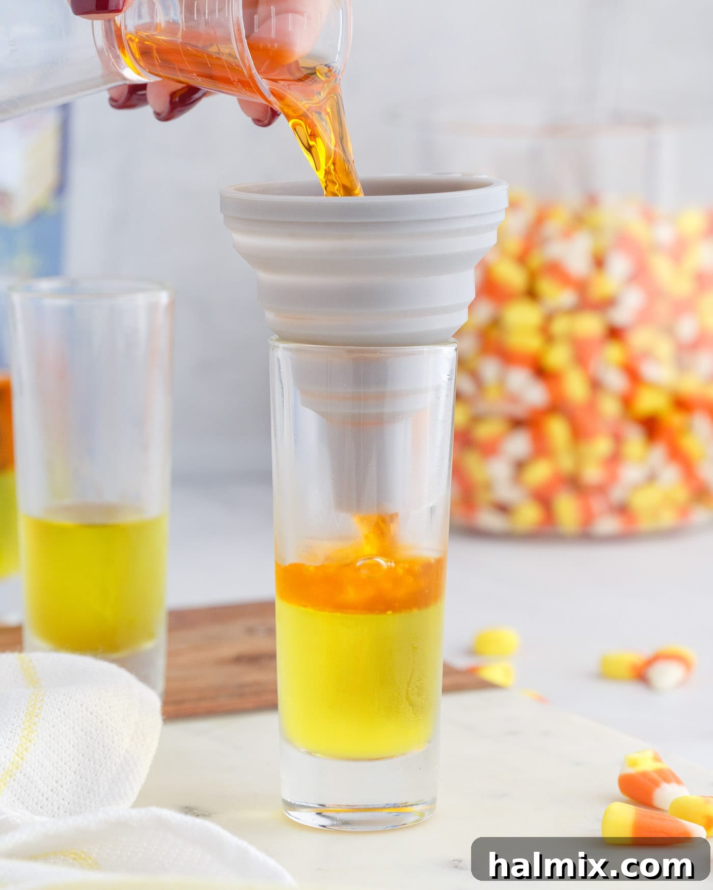 Pouring the orange jello mixture over the set lemon jello layer in a shot glass, creating the second vibrant layer of the candy corn jello shot.