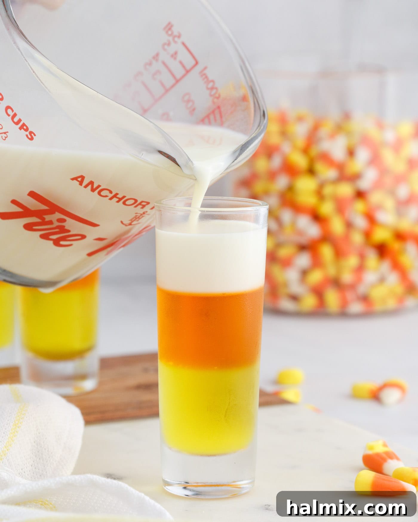 Carefully pouring the white condensed milk gelatin layer on top of the set orange and lemon layers in the shot glasses, completing the candy corn design.