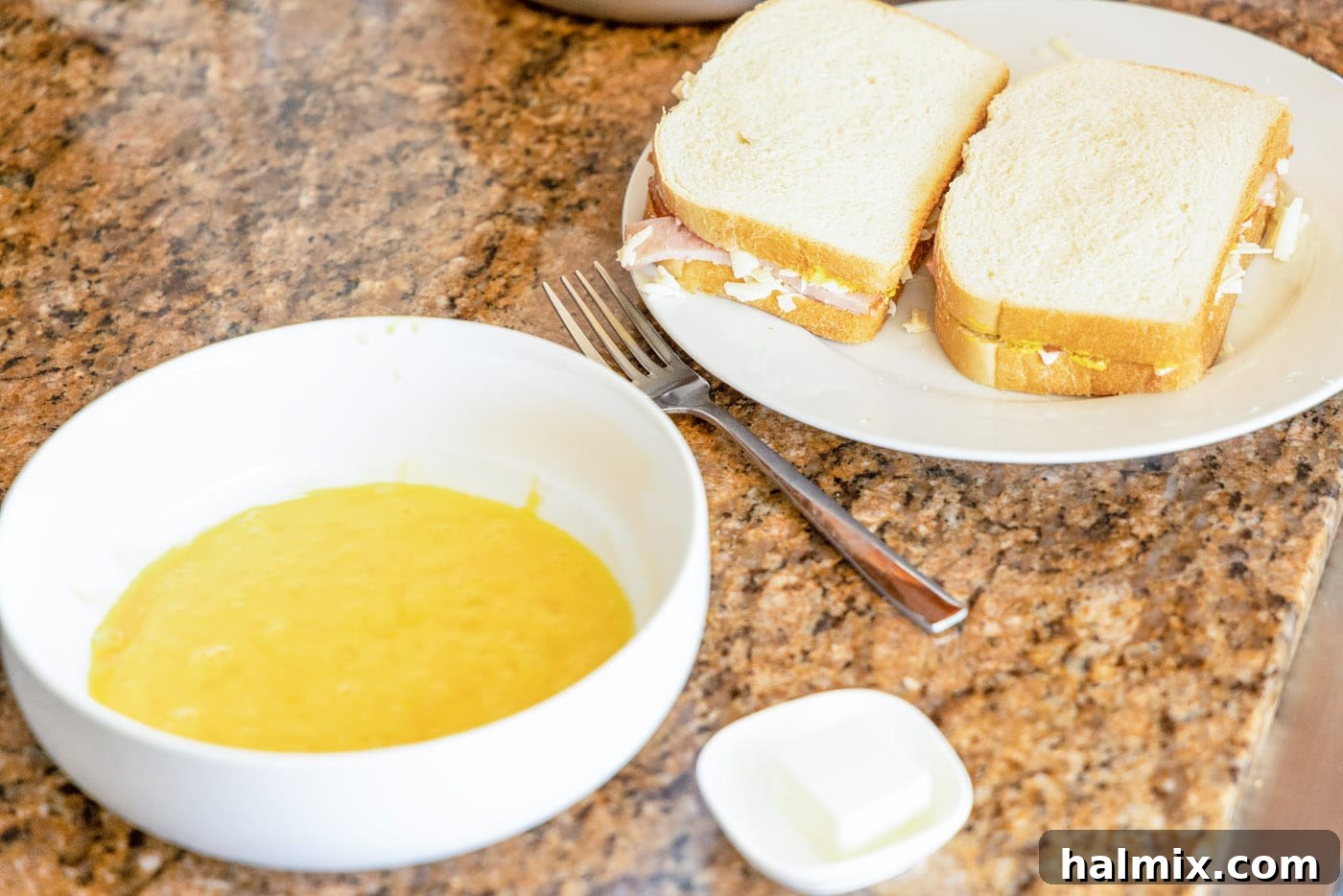 Whisked eggs in a shallow bowl, positioned next to an assembled Monte Cristo sandwich, prepared for dredging.