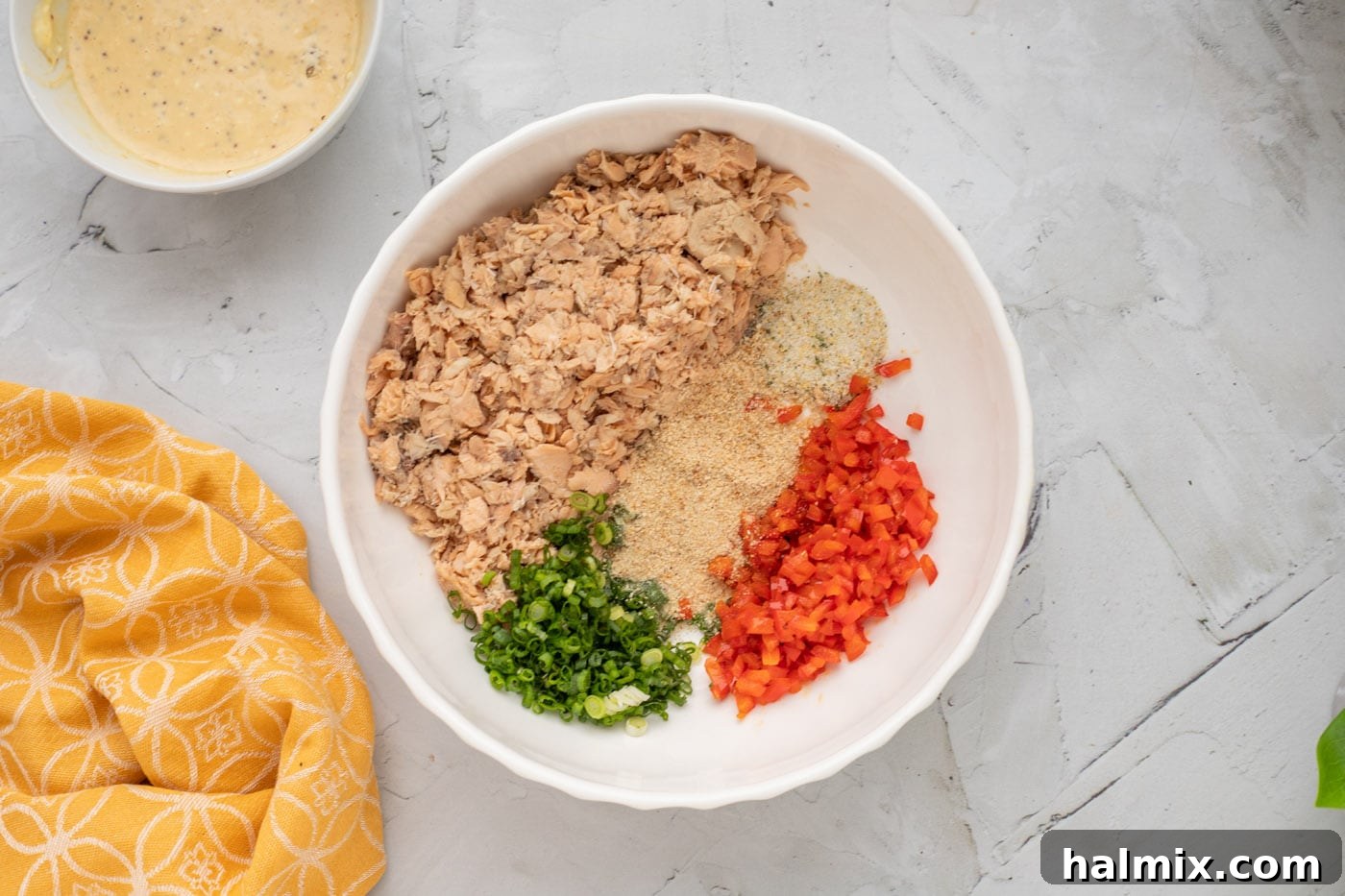 A large bowl filled with flaked salmon, green onion, diced bell pepper, and bread crumbs, waiting for the wet mixture.
