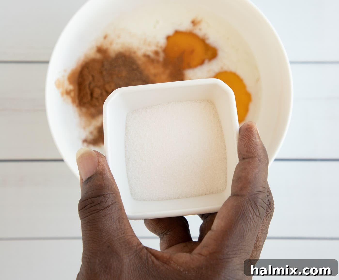 Hand holding sugar over bowl of spices and egg mixture, illustrating the mixing process for French Toast custard.