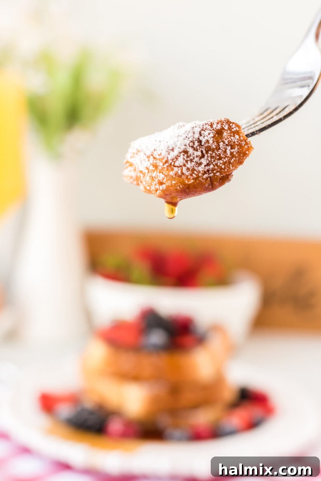 A single piece of French Toast on a fork, showing its fluffy texture, with a blurred background of a plate of French toast and toppings.