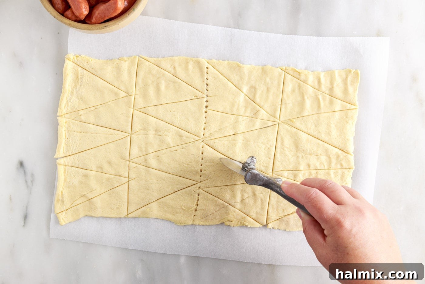 Sausage Swaddles 5 Slicing crescent roll dough into small triangles using a pizza cutter on a cutting board.