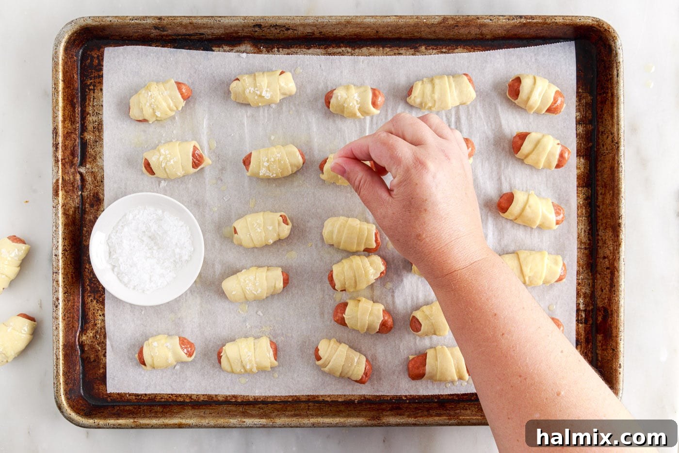 Sausage Swaddles 10 Hand sprinkling flaked sea salt over buttered pigs in a blanket on a baking sheet.