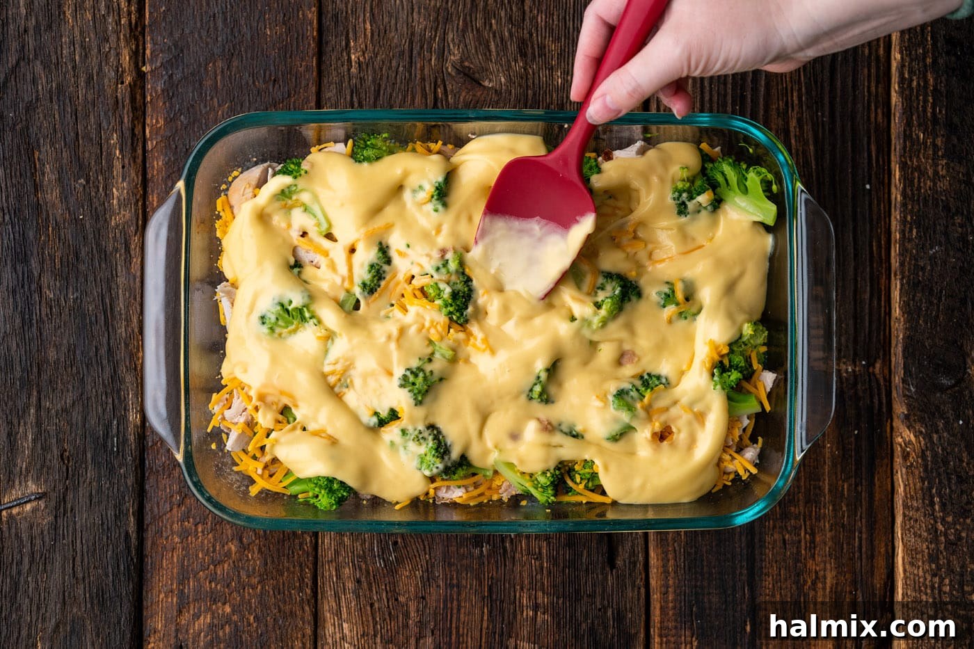 A spatula spreading cream of chicken soup smoothly over the layers of broccoli, chicken, and cheese in the casserole dish.