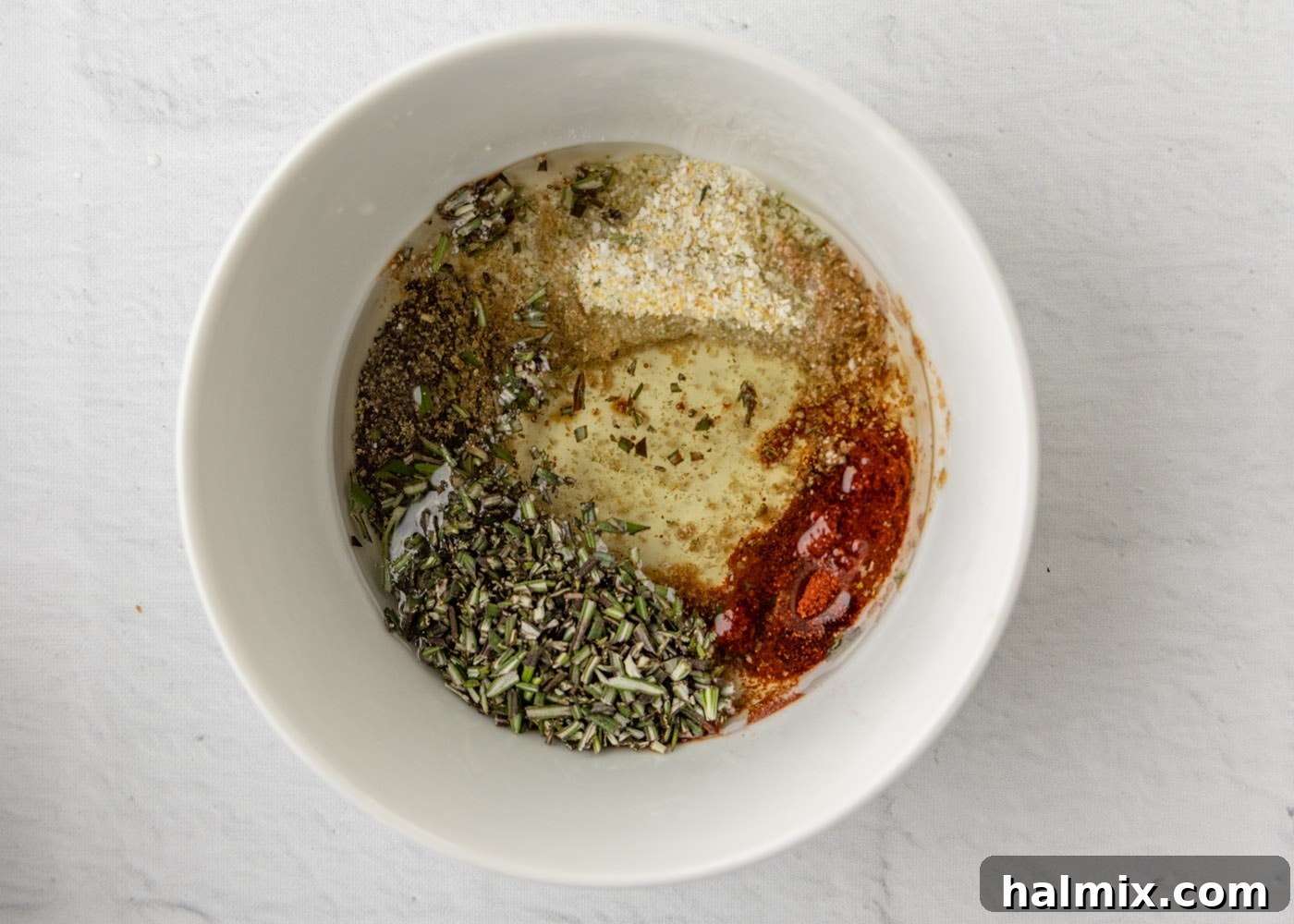 A bowl containing the mixed marinade ingredients: garlic salt, pepper, rosemary, paprika, liquid smoke, and olive oil