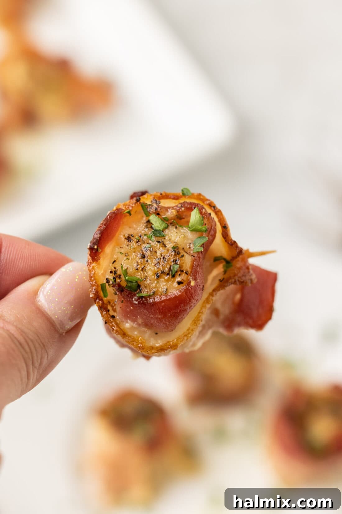 A hand holds a single perfectly cooked bacon wrapped scallop, showcasing its golden-brown exterior and crispy bacon.