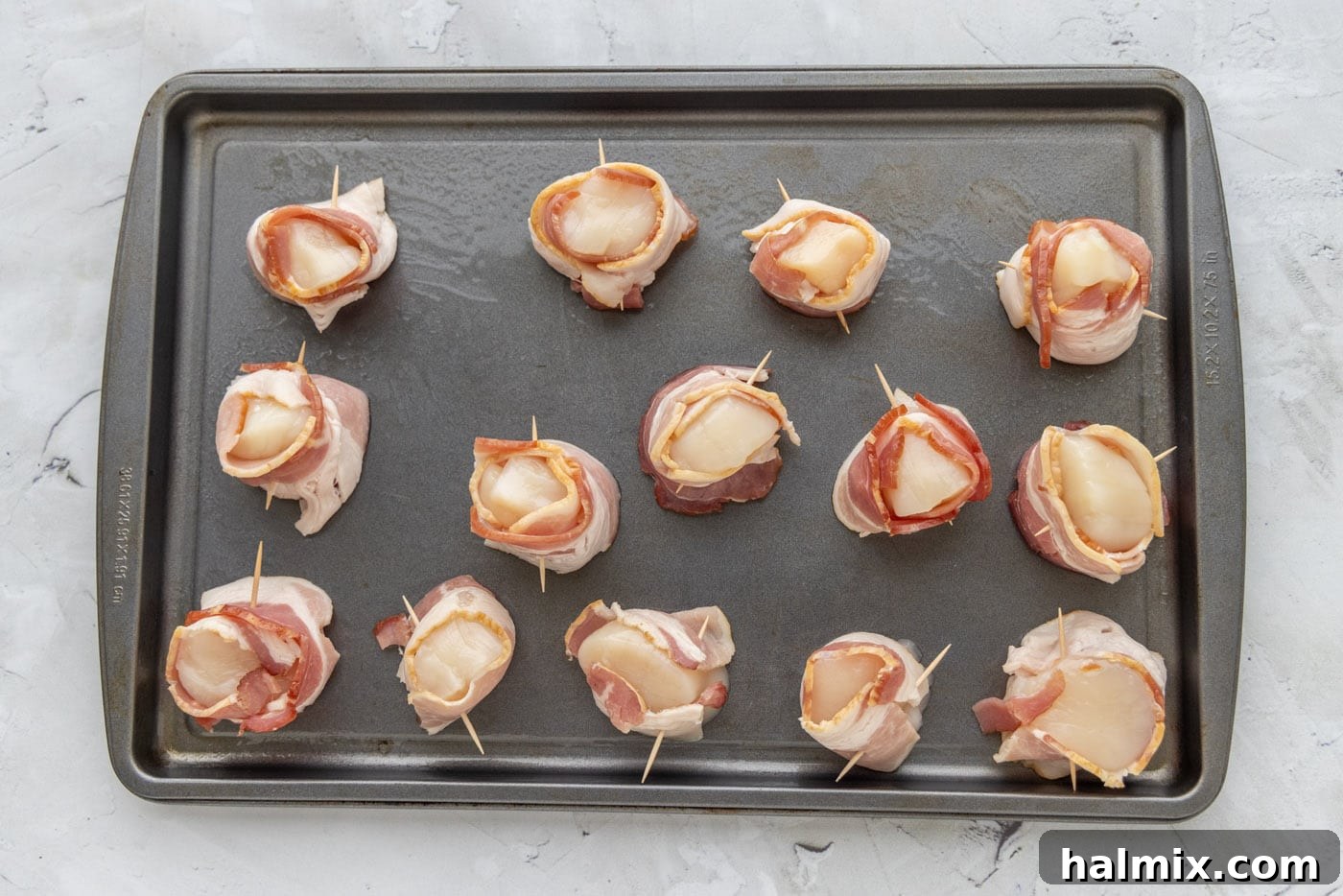 Raw sea scallops meticulously wrapped in bacon and secured with toothpicks, laid out on a baking sheet before cooking.