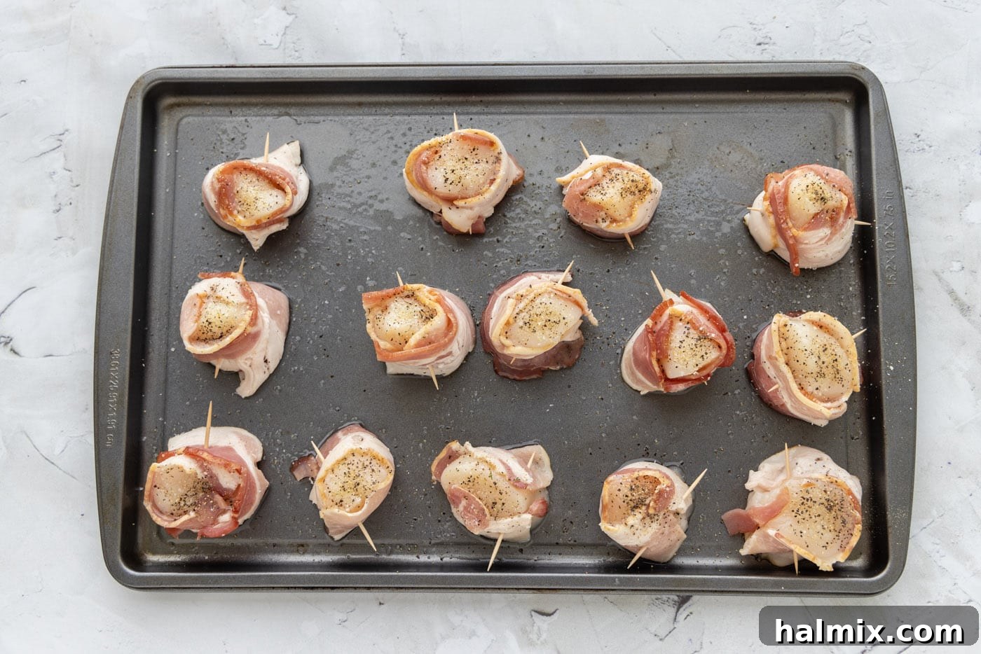 Bacon wrapped scallops on a baking sheet, lightly seasoned with olive oil, garlic salt, and pepper, glistening before broiling.