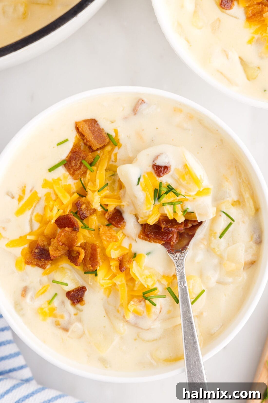 Hearty Loaded Baked Potato Soup 2 A spoonful of creamy Baked Potato Soup garnished with bacon, chives, and cheddar cheese, held over a rustic bowl of the soup.