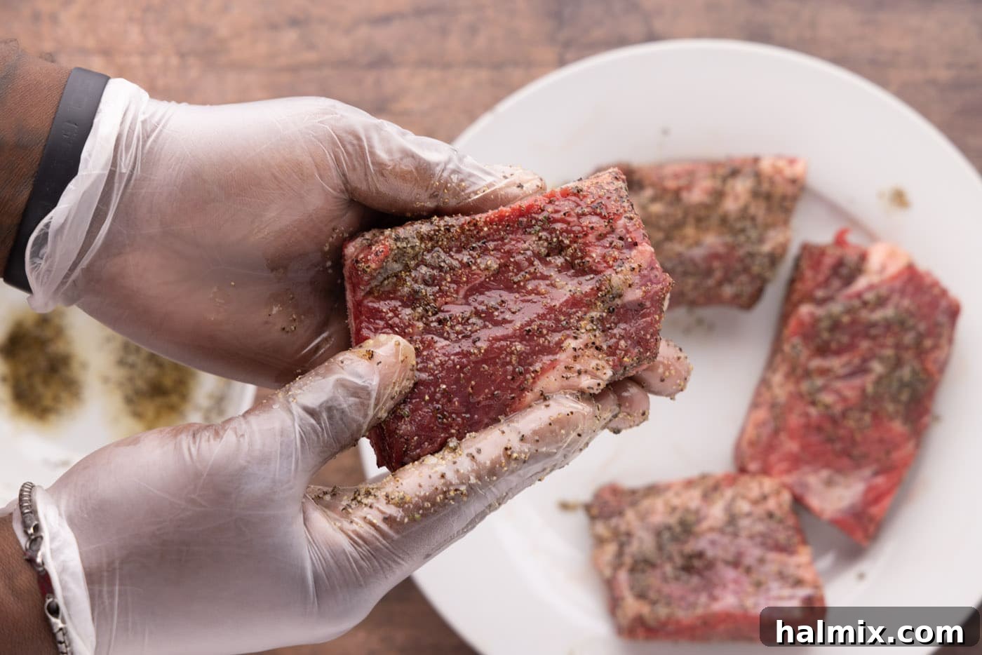 Fall-Off-The-Bone Beef Ribs 6 Gloved hands rubbing a mixture of pepper and garlic salt onto beef short ribs on a cutting board.