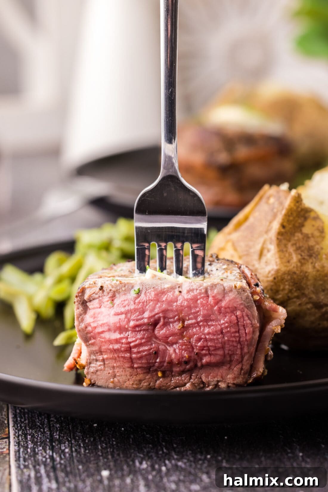Close up of a fork slicing into a perfectly cooked Bacon Wrapped Filet Mignon, showcasing its tender, pink interior and crispy, golden-brown bacon exterior.