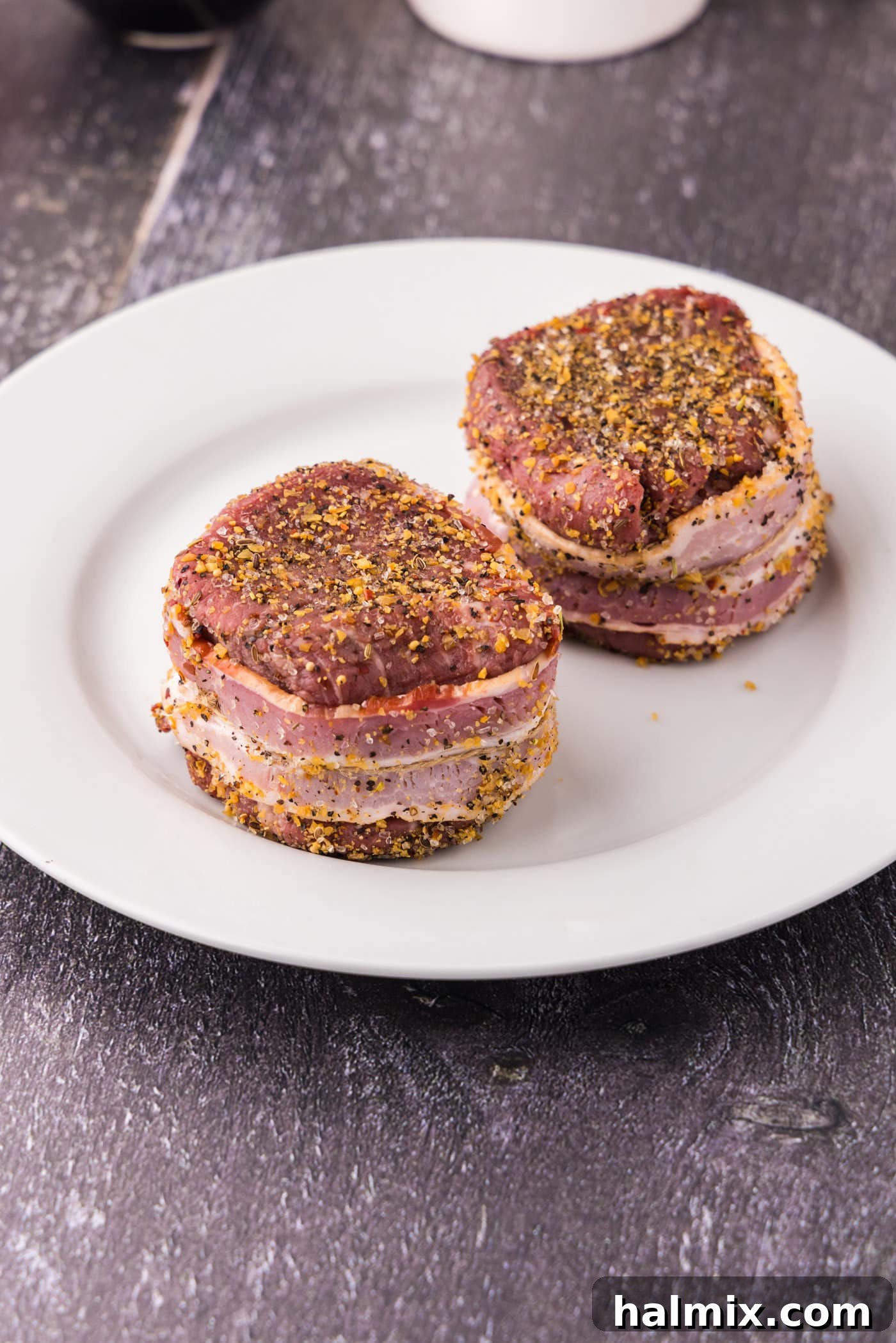 Bacon expertly wrapped around a raw filet mignon steak and secured with kitchen twine on a plate, ready for seasoning.