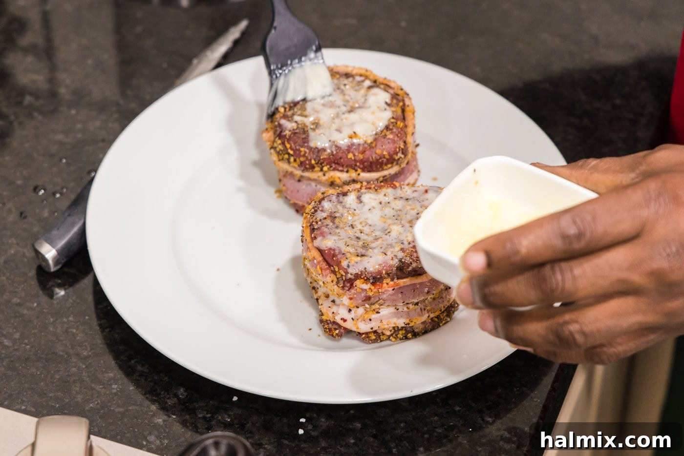 A hand using a pastry brush to apply softened butter to the top surface of a bacon-wrapped filet mignon.